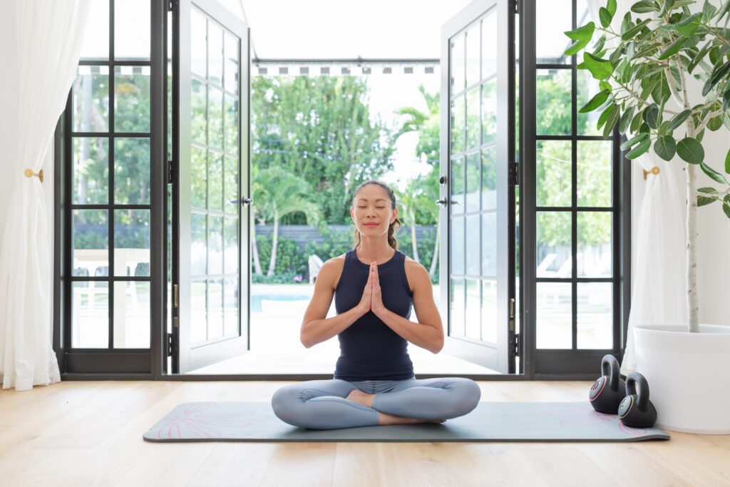 Woman meditating indoors with large black-framed glass doors opening to a poolside view